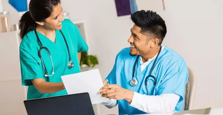 A female medical personnel smiling with a male medical personnel who has a single white piece of paper in his hand.