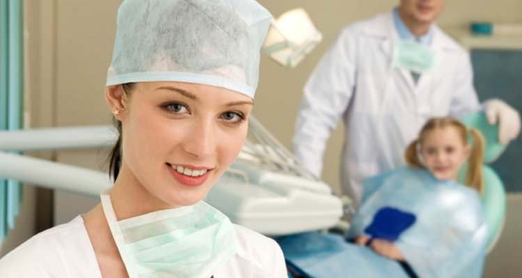 A dental assistant in full surgical gown and face mask hung around her neck in the foreground, a little girl who is a dental patient being administered dental surgery by another dentist.