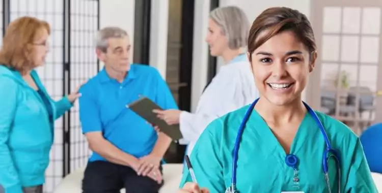 Female Medical Assistant smiling in the foreground while an elder patient is receiving advice from his doctor in the background.