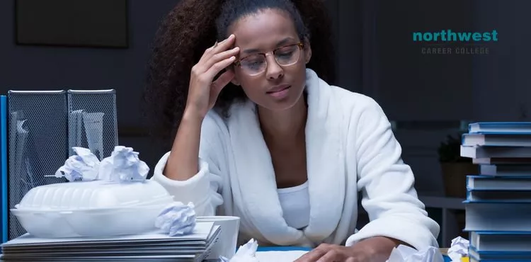A student in a robe looks stressed during Late-Night Study Sessions, surrounded by crumpled papers and stacks of books.