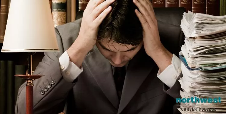 A paralegal is stressed, hand on his head, next to a stack of papers.