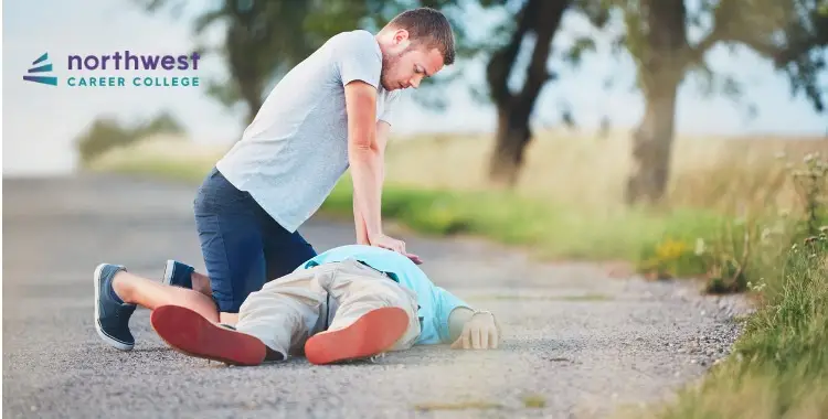 A person performs CPR on an unresponsive individual lying on a gravel road, with trees in the background.