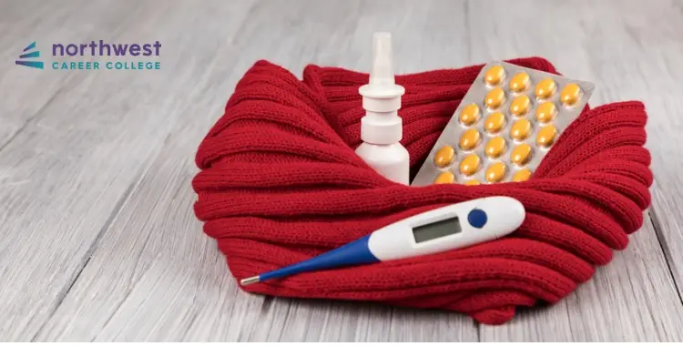 A red knitted bowl holds a thermometer, nasal spray, and a blister pack of yellow pills on a wooden surface.