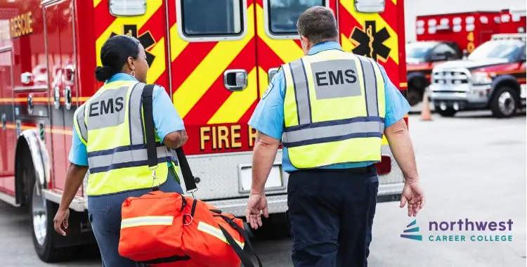 Two EMS workers in reflective vests walk towards an ambulance, one carrying a medical bag.
