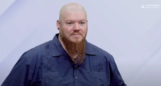 A bald man with a long beard stands in front of a light background, wearing a dark shirt with embroidery.
