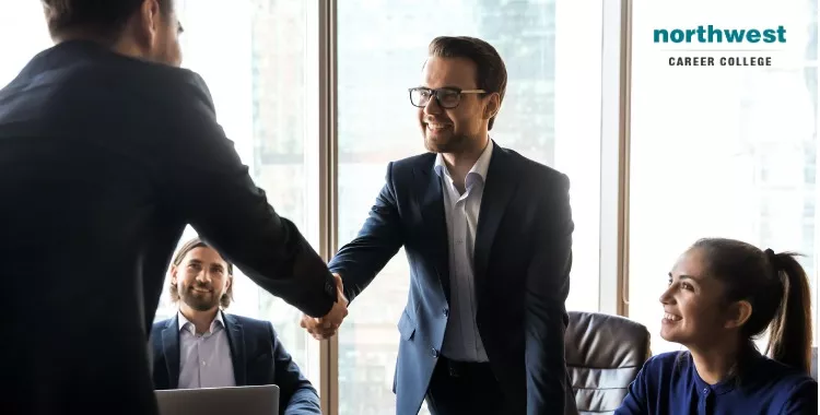 A professional meeting where an administrative assistant shakes hands with a colleague, while others observe.
