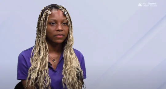 A young person with long, braided hair sits in front of a light background, wearing a purple shirt.