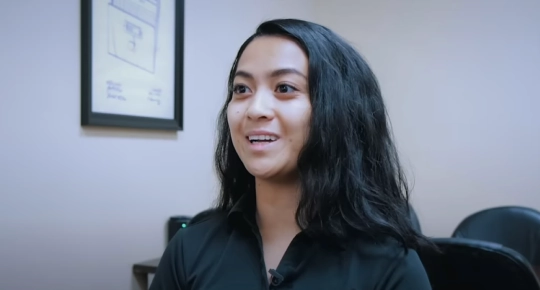 A young woman with long, dark hair smiles while speaking in an office setting. A framed drawing is visible in the background.
