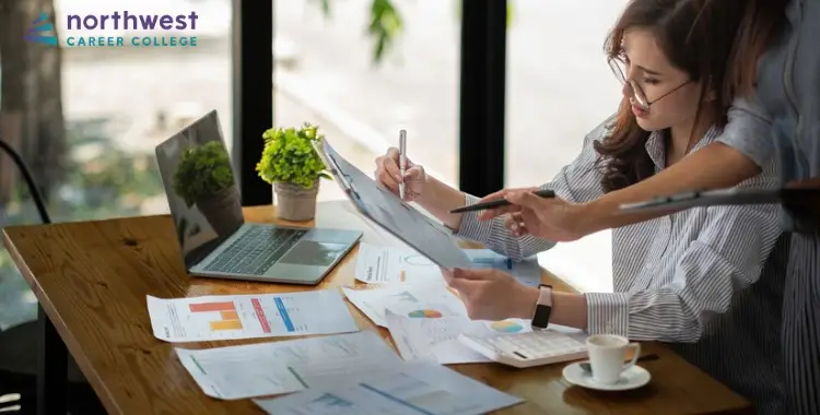 A woman analyzes financial management reports at a table with a laptop and documents, collaborating with another person.