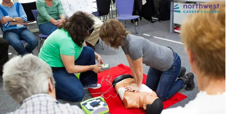 Two women perform CPR training on a manikin while others observe in a classroom setting.