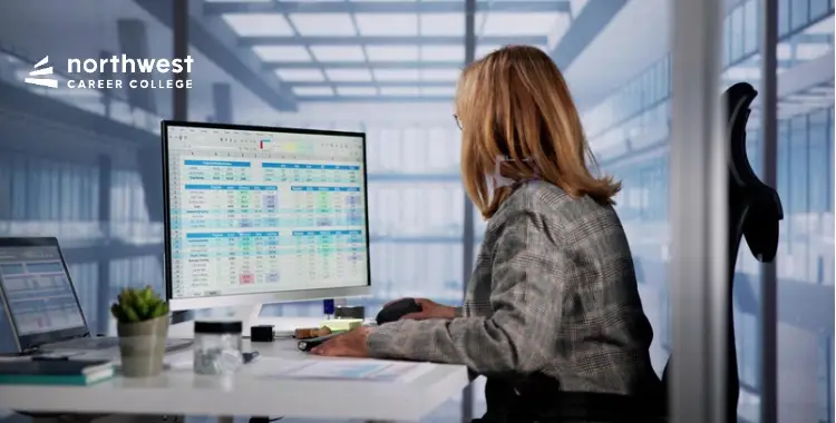 A person with long hair works at a desk, analyzing data on a large computer screen in a modern office setting.
