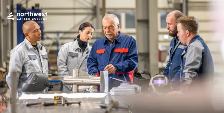 Five individuals in work attire gather around a table, discussing metalwork in a workshop setting.