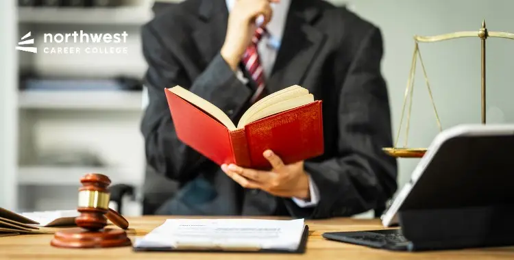 A suited individual holds a book, pondering, with a gavel and scales of justice on the desk.