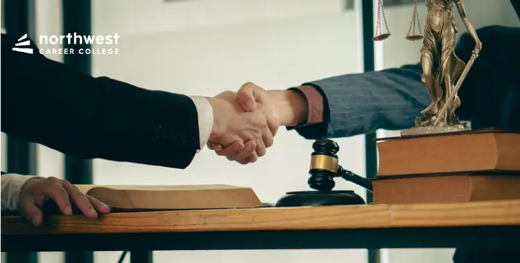 Two people shaking hands over a desk with legal books and a gavel, symbolizing agreement or partnership.