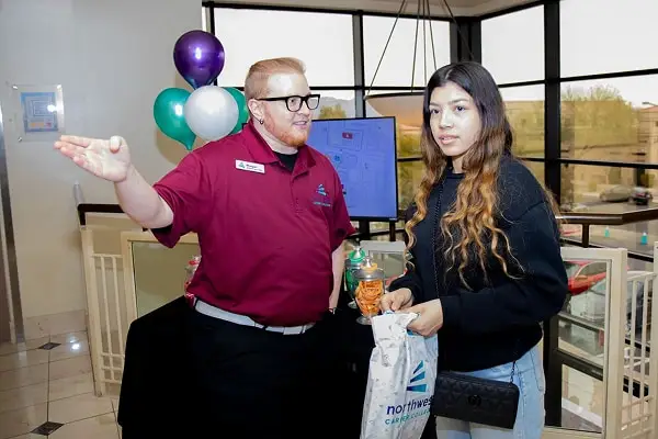 A man in a maroon shirt gestures while speaking to a woman holding a bag and drink in a bright, modern setting.