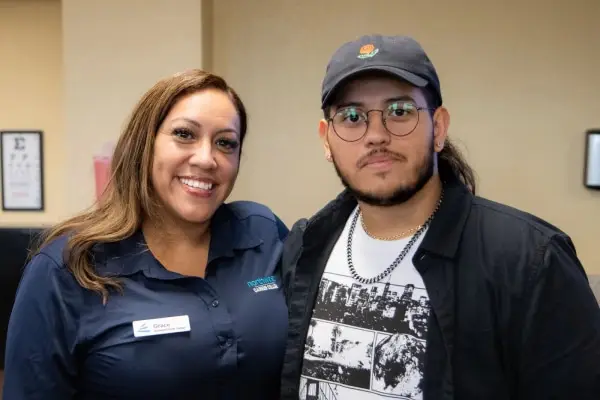 Two smiling individuals pose together in a casual setting, one wearing a name tag and the other a cap and glasses.