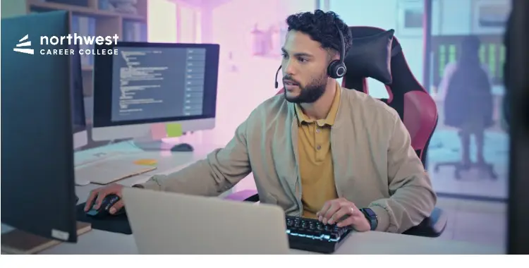 A man wearing a headset works at a computer desk with multiple monitors in a modern office setting.