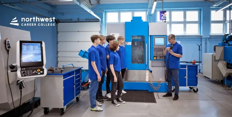 Students in blue shirts observe an instructor by a CNC machine in a workshop setting.