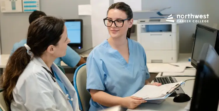 Two healthcare students in scrubs engage in conversation at a computer station, with paperwork and equipment around them.