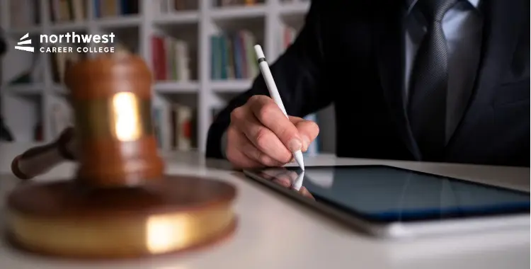 A person in a suit writes on a tablet beside a gavel, with bookshelves in the background.