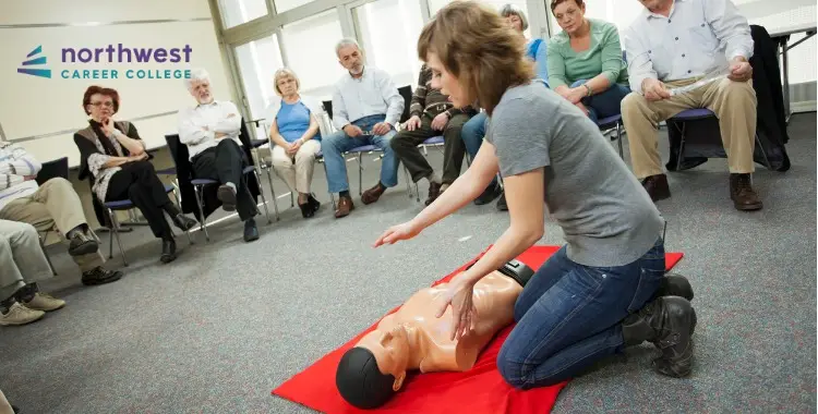 A woman demonstrates CPR on a mannequin while an audience observes in a classroom setting.