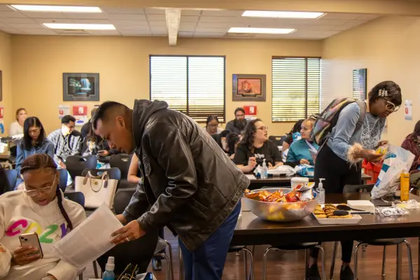 A diverse group of people seated in a classroom, some eating snacks, others on their phones, engaged in conversation.