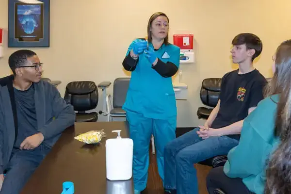 A healthcare professional in scrubs demonstrates a procedure to three seated teenagers in a clinical setting.