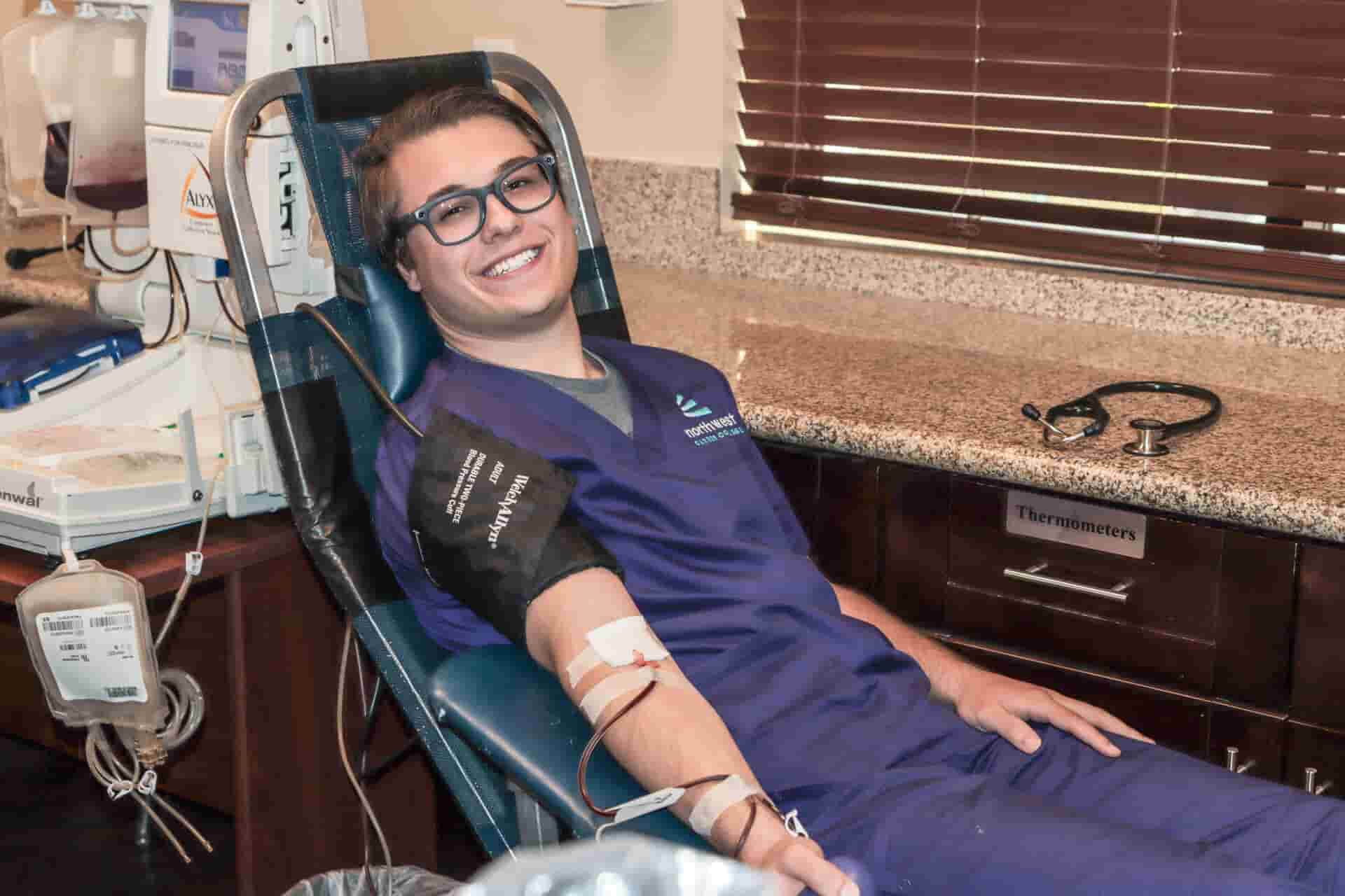 A smiling young man in scrubs sits in a medical chair with an IV in his arm, surrounded by medical equipment.