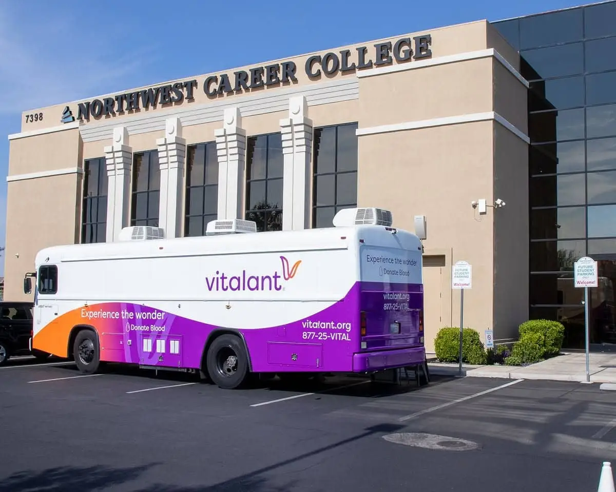 A blood donation bus parked outside Northwest Career College, featuring the Vitalant logo and donation information.
