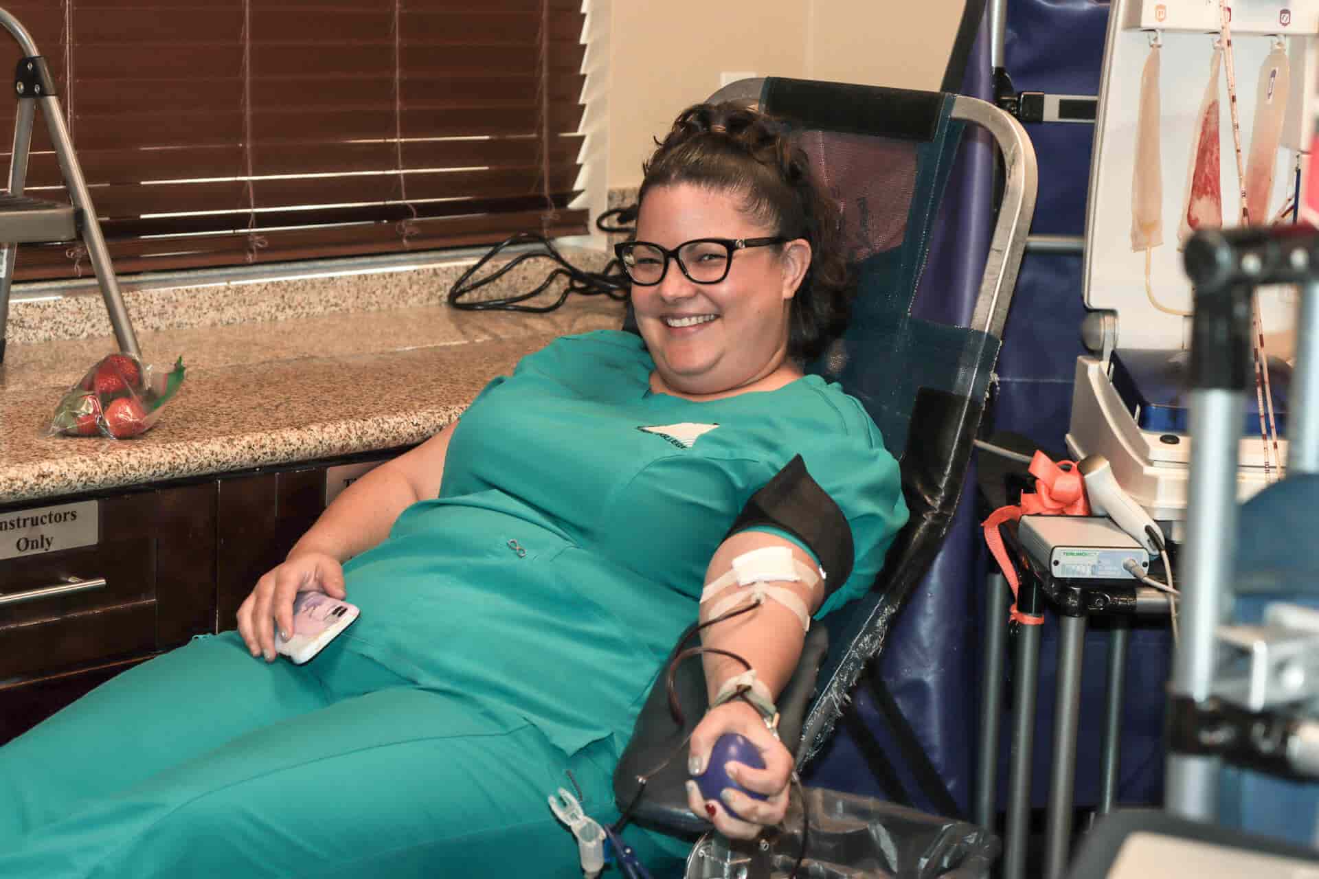 A smiling woman in scrubs donates blood while holding a stress ball in a medical setting.