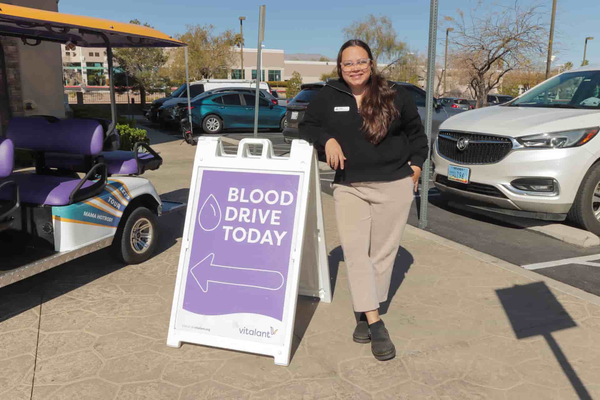 A woman stands next to a sign promoting a blood drive, with cars and a golf cart in the background.