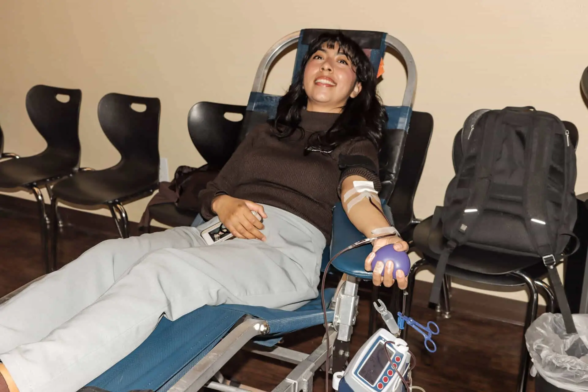 A smiling person donates blood while seated, with medical equipment and chairs in the background.