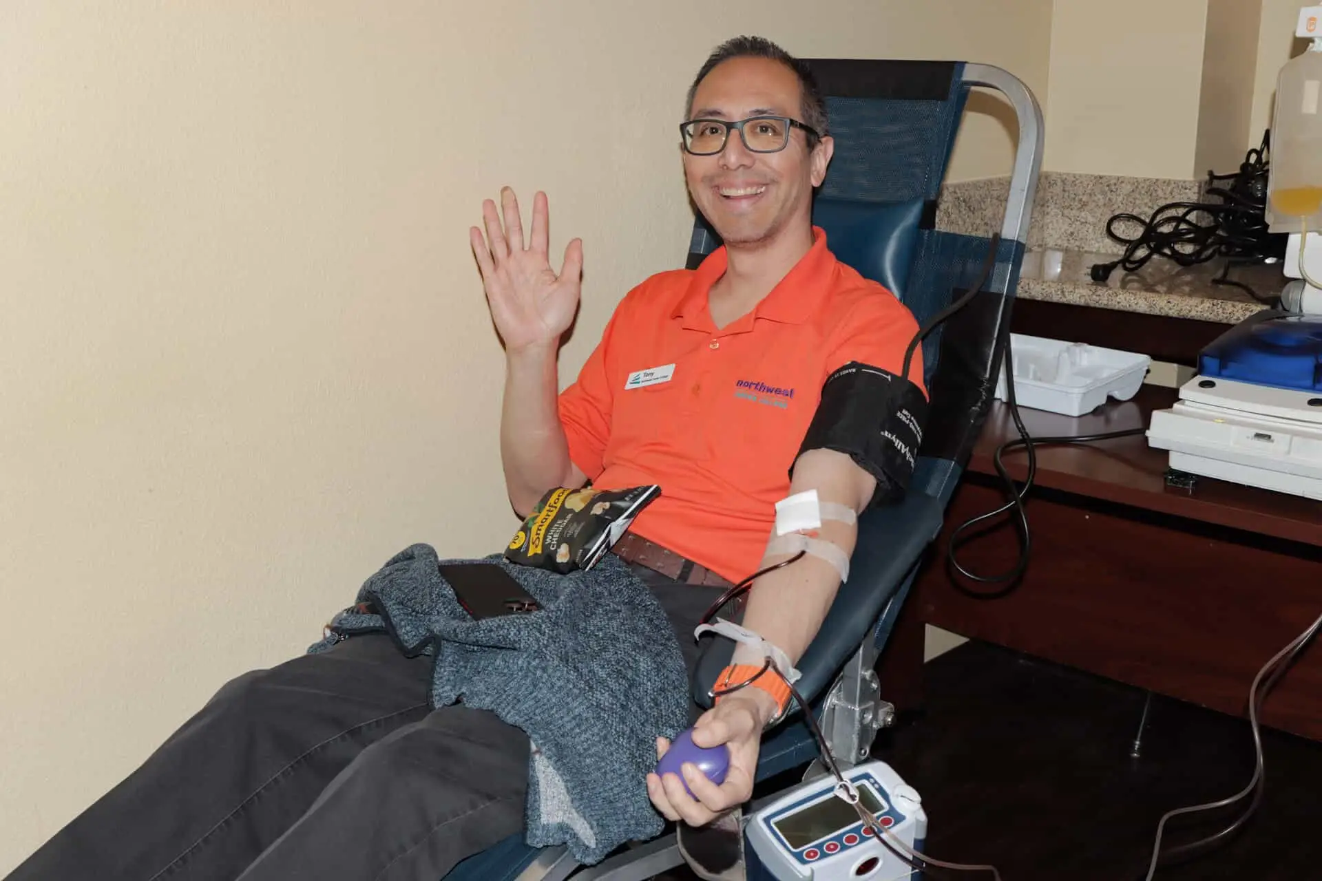 A smiling man in an orange shirt sits in a donation chair, waving with one hand while the other is connected to a blood.
