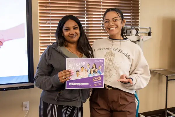 Two women hold a Gift Card Raffle sign, smiling in a well-lit room with a presentation screen in the background.