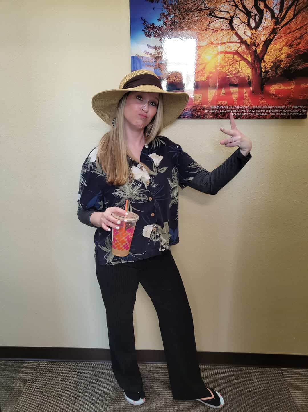 Woman in a floral shirt and wide-brimmed hat poses playfully with a drink in front of a wall featuring a nature-themed.