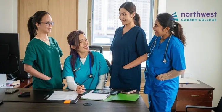 Four healthcare students in scrubs engage in discussion around a desk, smiling and collaborating in a bright classroom.