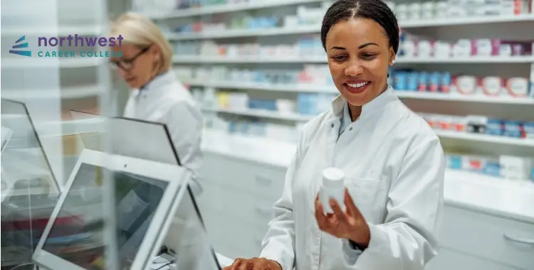 A pharmacist smiles while examining a medication bottle, with another pharmacist in the background at a pharmacy.