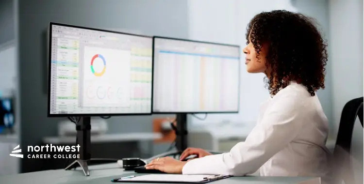 A woman in a white lab coat works at a desk with dual monitors displaying data and charts.