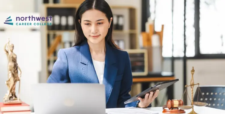 A woman in a blue blazer works on a laptop, holding a tablet, with legal symbols in the background.