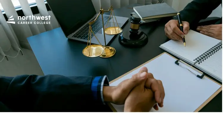 Two people in business attire sit at a desk with a laptop, scales of justice, and a notebook, discussing legal matters.