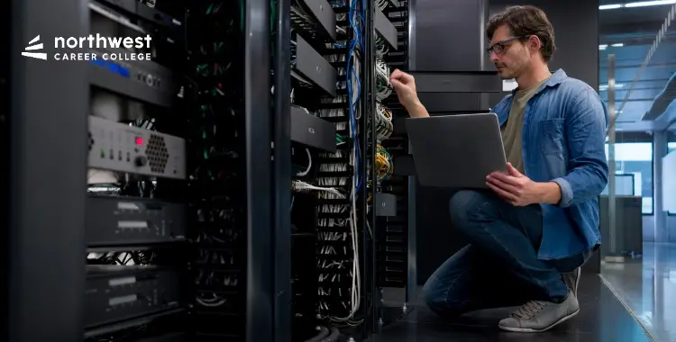 A man kneels beside server racks, using a laptop to troubleshoot or manage network connections.