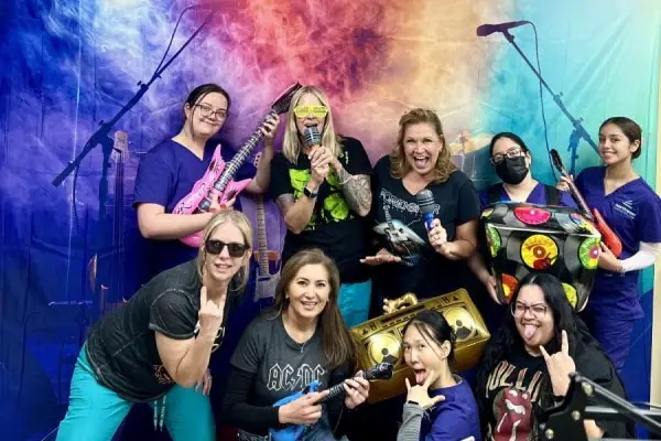 A group of women in colorful outfits pose with musical instruments and props, celebrating a fun, rock-themed event.