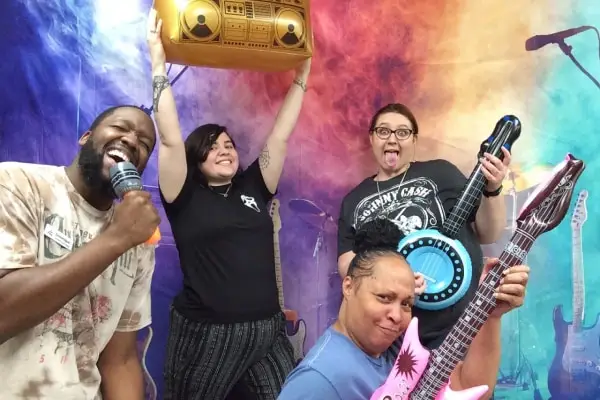 Four friends pose playfully with musical props, including a microphone, guitar, and boombox, against a colorful backdrop.