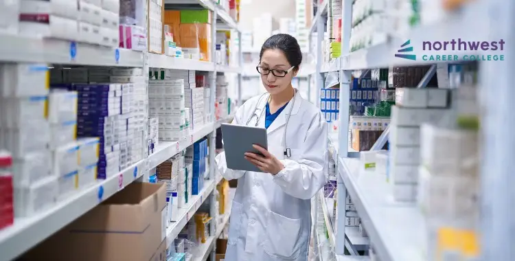 A pharmacist in a lab coat uses a tablet while standing in a pharmacy aisle filled with medication boxes.