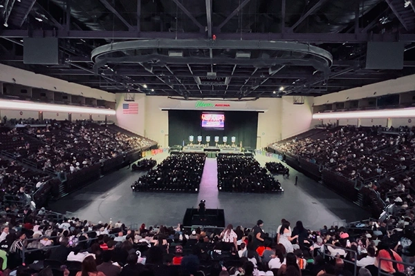 A large crowd gathers in an arena for a graduation ceremony, with a stage at the front and attendees in black robes.