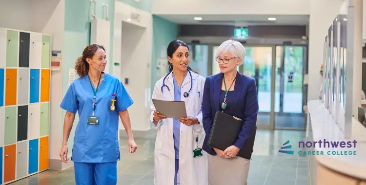 Three women in a healthcare setting discuss documents while walking, wearing scrubs and professional attire.