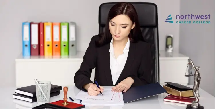 A woman in a suit sits at a desk, writing on a document with books and folders in the background.