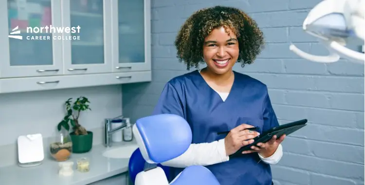 Smiling dental professional in scrubs holding a tablet in a modern dental office.