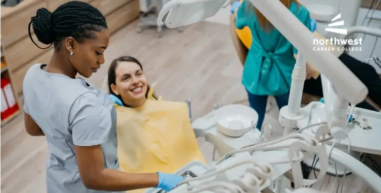 A dental assistant works on a patient in a clinic, while another staff member observes in the background.