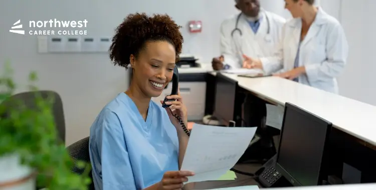 A smiling woman in scrubs talks on the phone while reviewing documents at a reception desk in a medical setting.